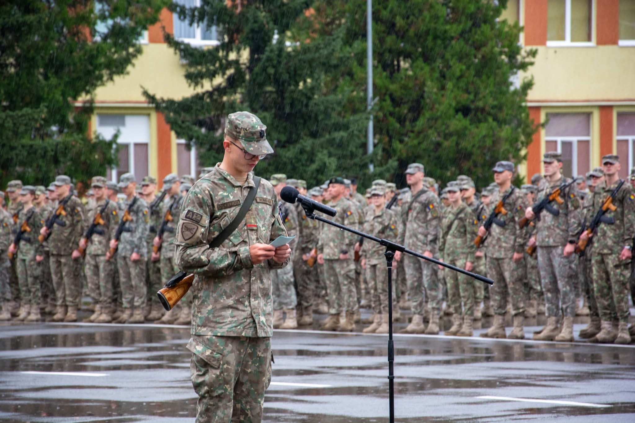 Soldat militar în ceremonie de absolvire în uniforme de camuflaj în fața unui grup de militari în linie, în parcul orașului Sibiu, România.
