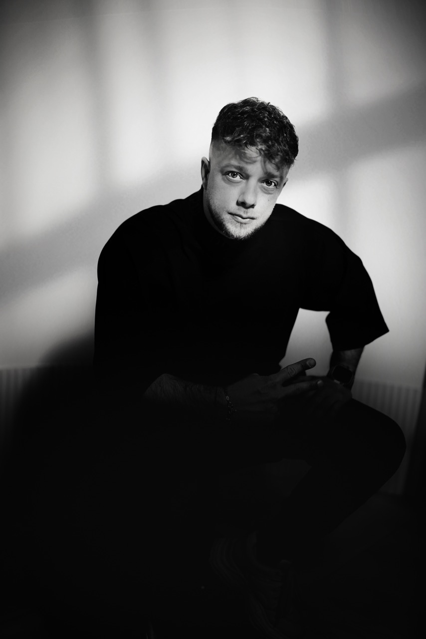 Tânăr bărbat într-un portret negru și alb, cu expresie serioasă, purtând un pulover negru, foto artistică, iluminare dramatică pentru conținut vizual de înaltă calitate.