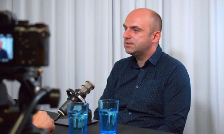 Barbat vorbind la interviu pentru Ora de Sibiu, în interior, cu două pahare cu apa pe masă, în timpul unei discuții sau conferințe de presă.