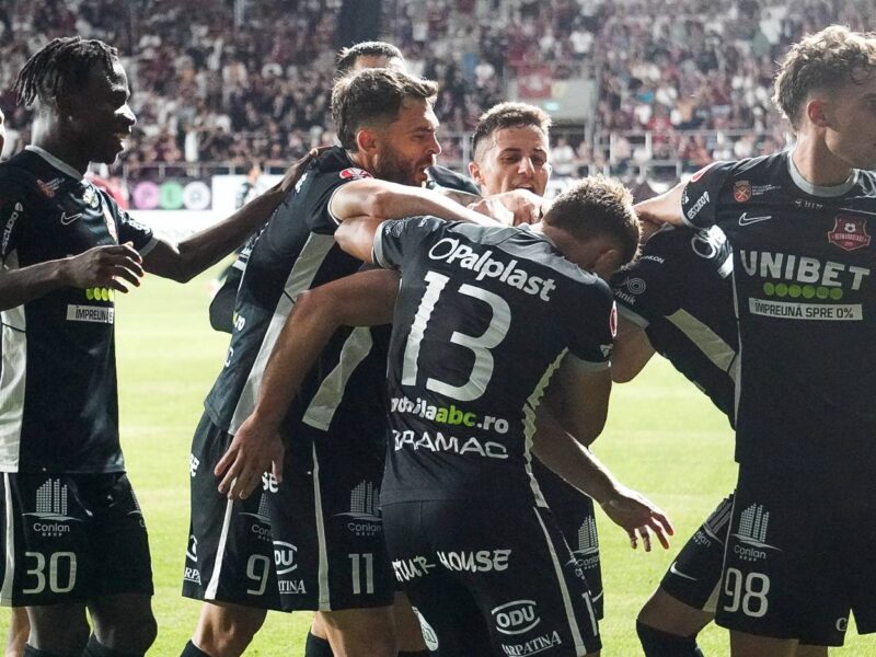 Jucători de fotbal în echipament negru, celebrați pe teren în timpul unui meci, cu mulțumiri și bucurie, în centrul atenției sportului profesionist, promovând echipa și spiritul competiției dinamice.