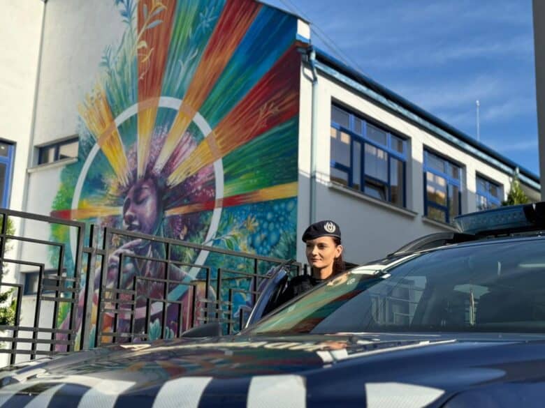 Artiștă de poliție alături de mașina de poliție în fața unui mural colorat pe o clădire din Sibiu, reprezentând o figură feminină cu un cerc de culori vibrant în jurul capului, pe stradă.
