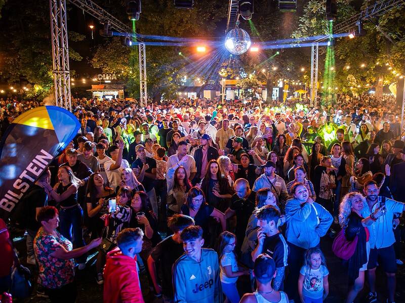 Mulțime de oameni la un concert în aer liber, seara, sub lumini colorate și glob disco