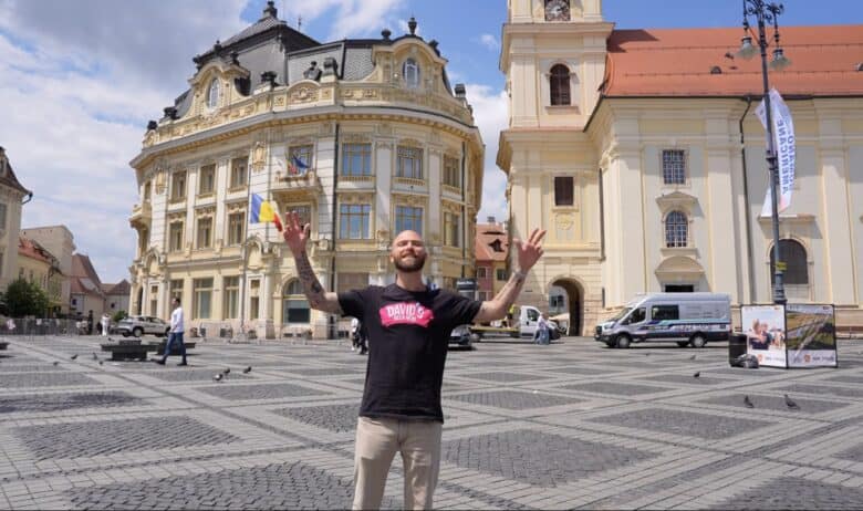 Vloggerul american David Hoffmann, cu brațele deschise, în Piața Mare din Sibiu, în fața Primăriei și a Bisericii Iezuiților – captură din episodul Davidsbeenhere despre Sibiu
