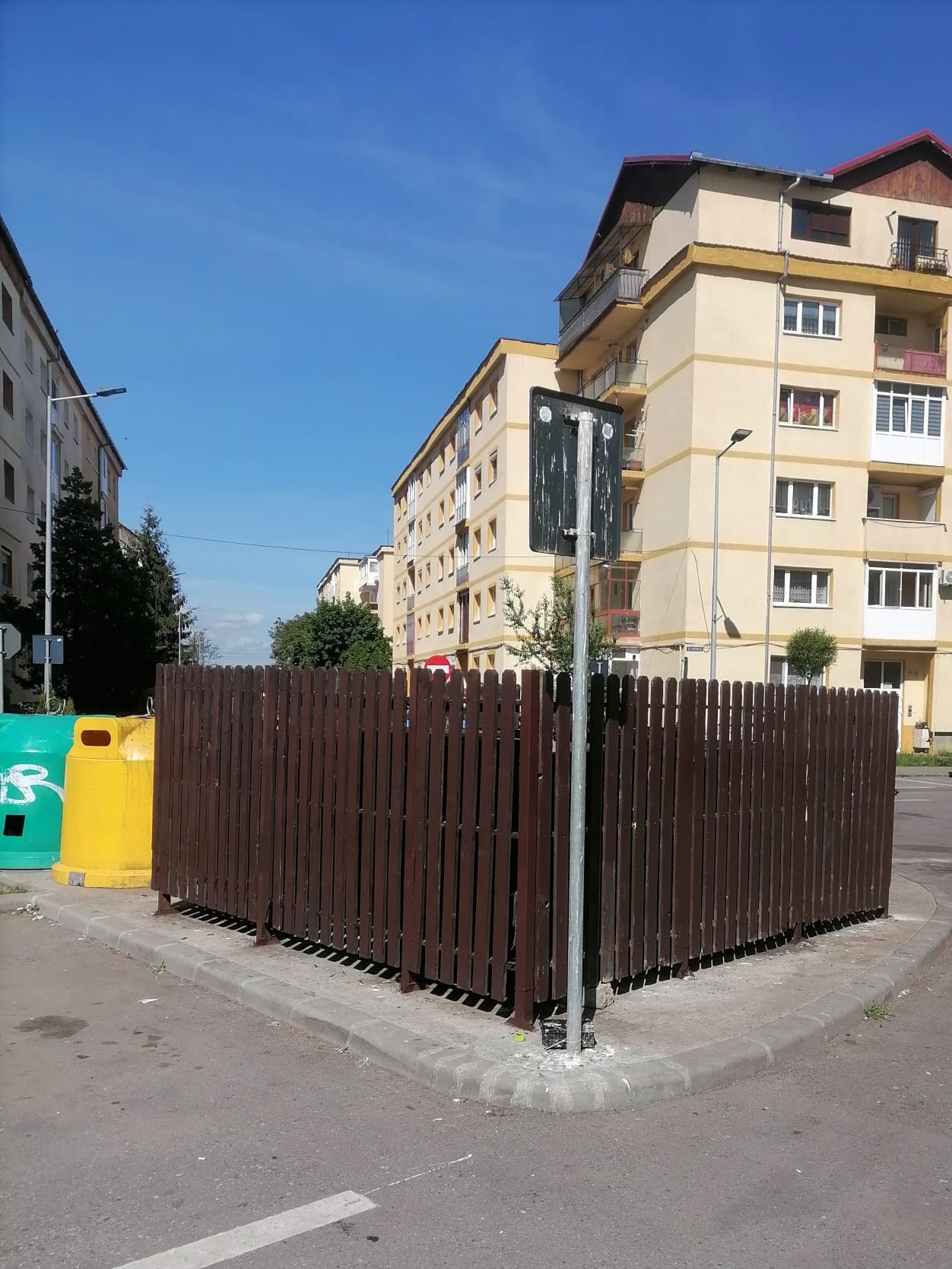 Platformă de gunoi îngrădită, între blocuri de locuințe din Sibiu, cu container albastru pentru hârtie și carton lângă trotuar