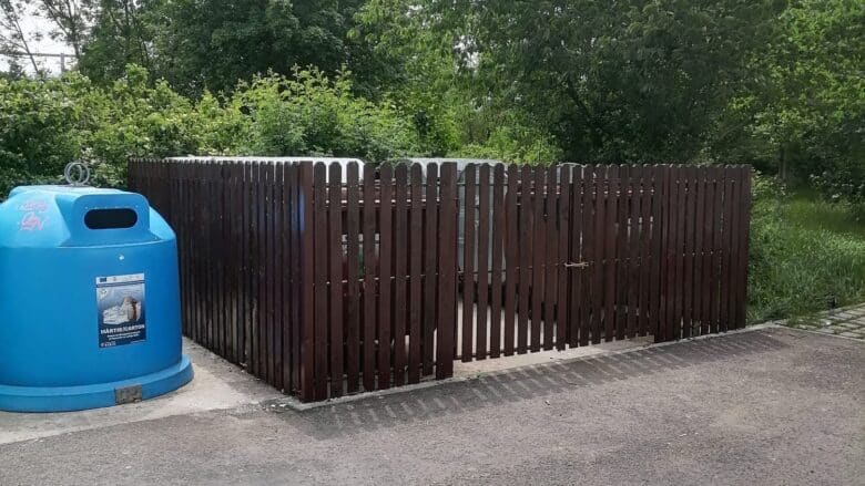 Platformă de colectare a deșeurilor din Sibiu, împrejmuită cu gard din lemn maro, cu un container albastru pentru hârtie și carton în față