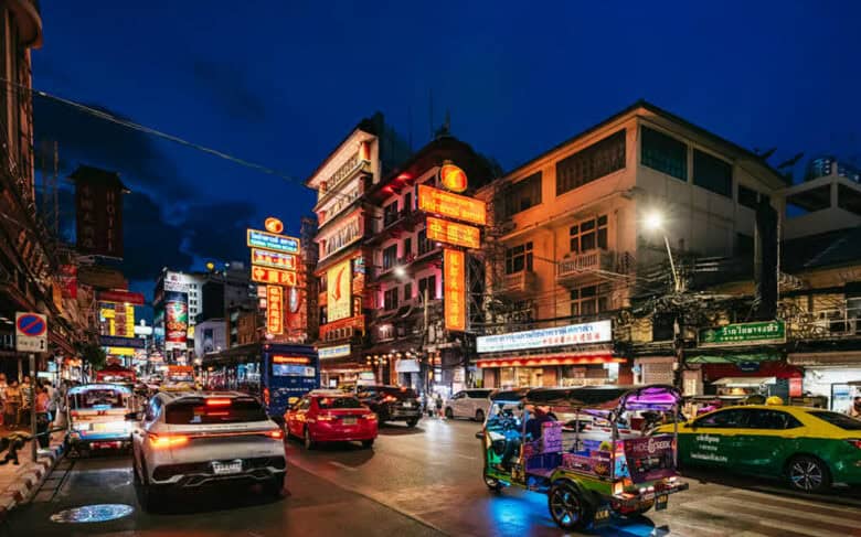 Stradă aglomerată din Bangkok noaptea, cu reclame neon, tuk-tuk-uri și taxiuri colorate